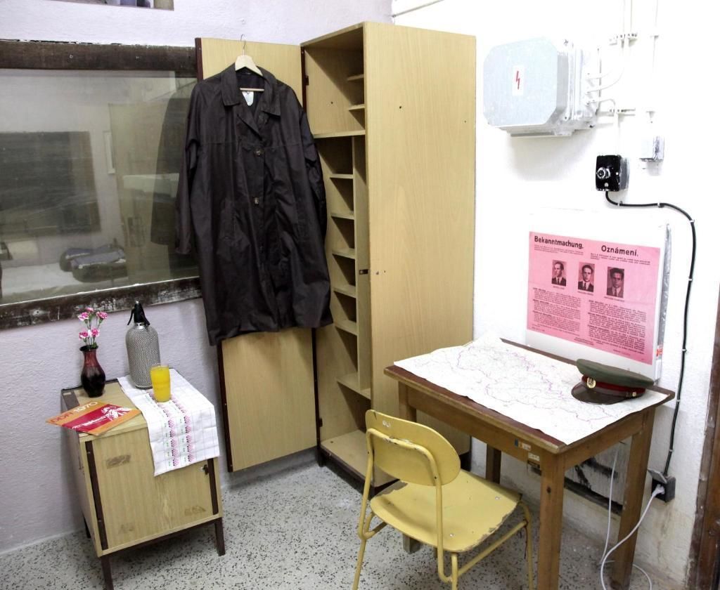A small office with a wooden desk, a chair, a coat rack, a small table with a bottle and a glass, and a poster on the wall.