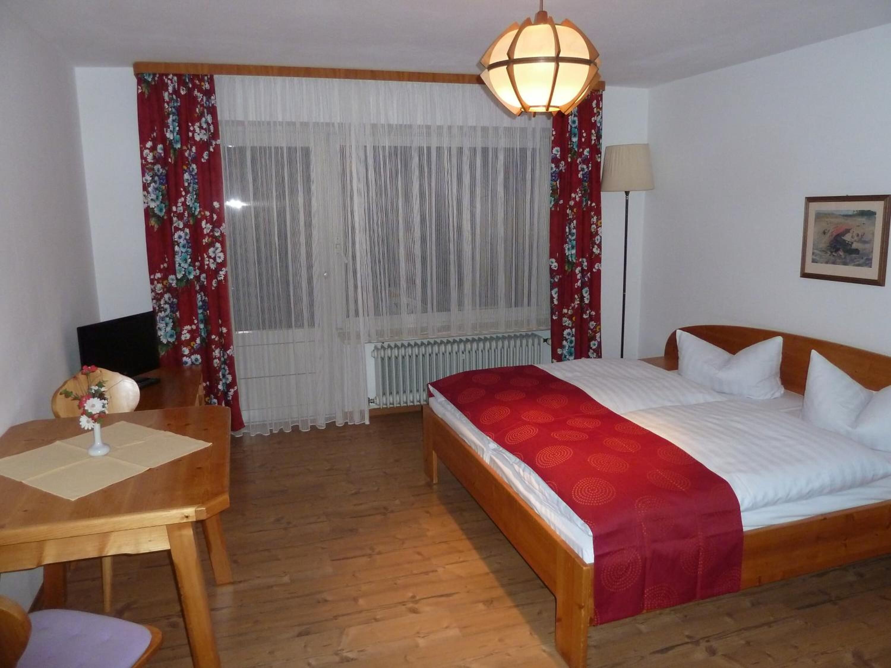A hotel room with a wooden bed, a desk with a chair, a radiator, a lamp, and a window with curtains.