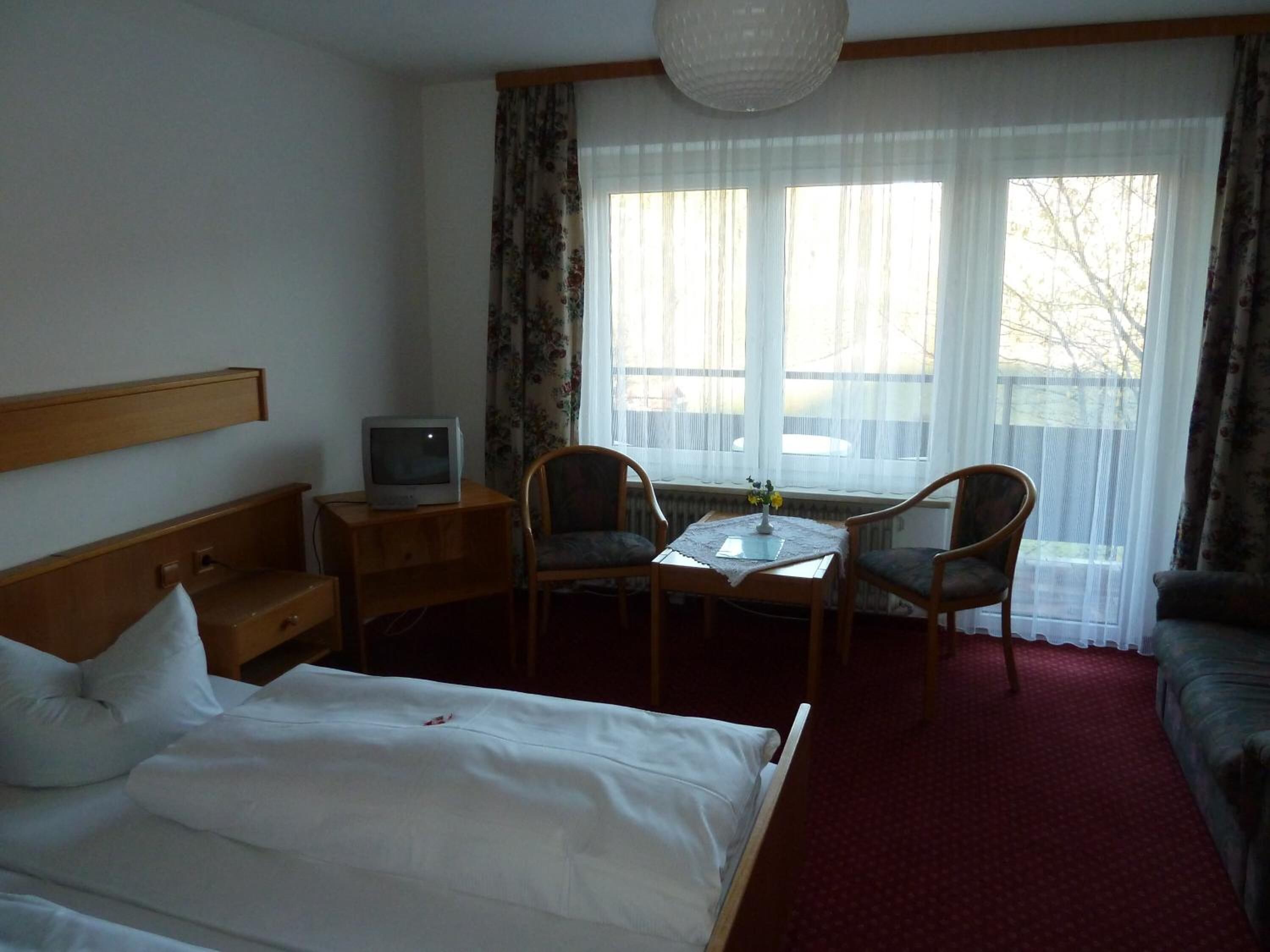 Habitación de hotel con una cama, un escritorio con televisor, dos sillas, una mesita de noche con un florero y una ventana con cortinas.