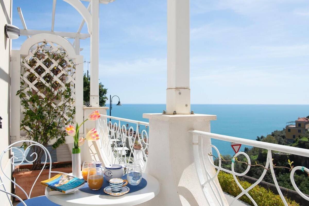 Un balcon avec une balustrade blanche, une table avec un livre, un vase rempli de fleurs et une vue sur la mer.