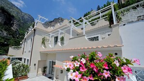 Un bâtiment à deux étages doté d’un balcon, d’une grande plante en pot aux fleurs roses et ayant une chaîne de montagnes en arrière-plan.