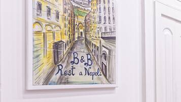 Une œuvre d’art encadrée représentant une scène de rue, avec l’inscription « B&B Rest a Napoli » sur un mur blanc, une table de petit-déjeuner avec du pain, des œufs et des viennoiseries, et une porte sur la droite.