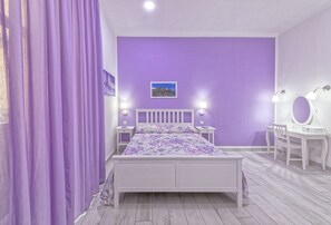 Un dormitorio con una pared morada, una cama blanca, un escritorio con un espejo y dos lámparas de noche.