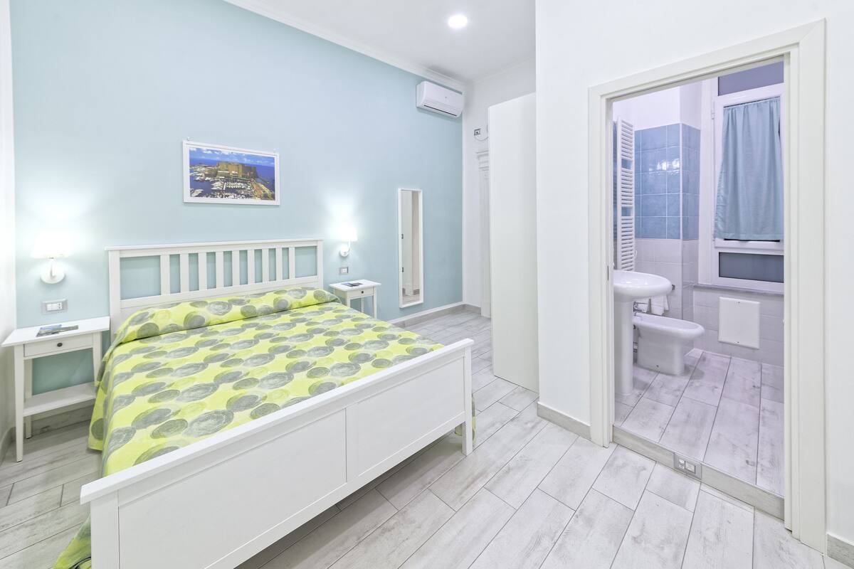 A bedroom with a bed, bedside tables, a nightstand, a mirror, and a bathroom with a toilet and sink.