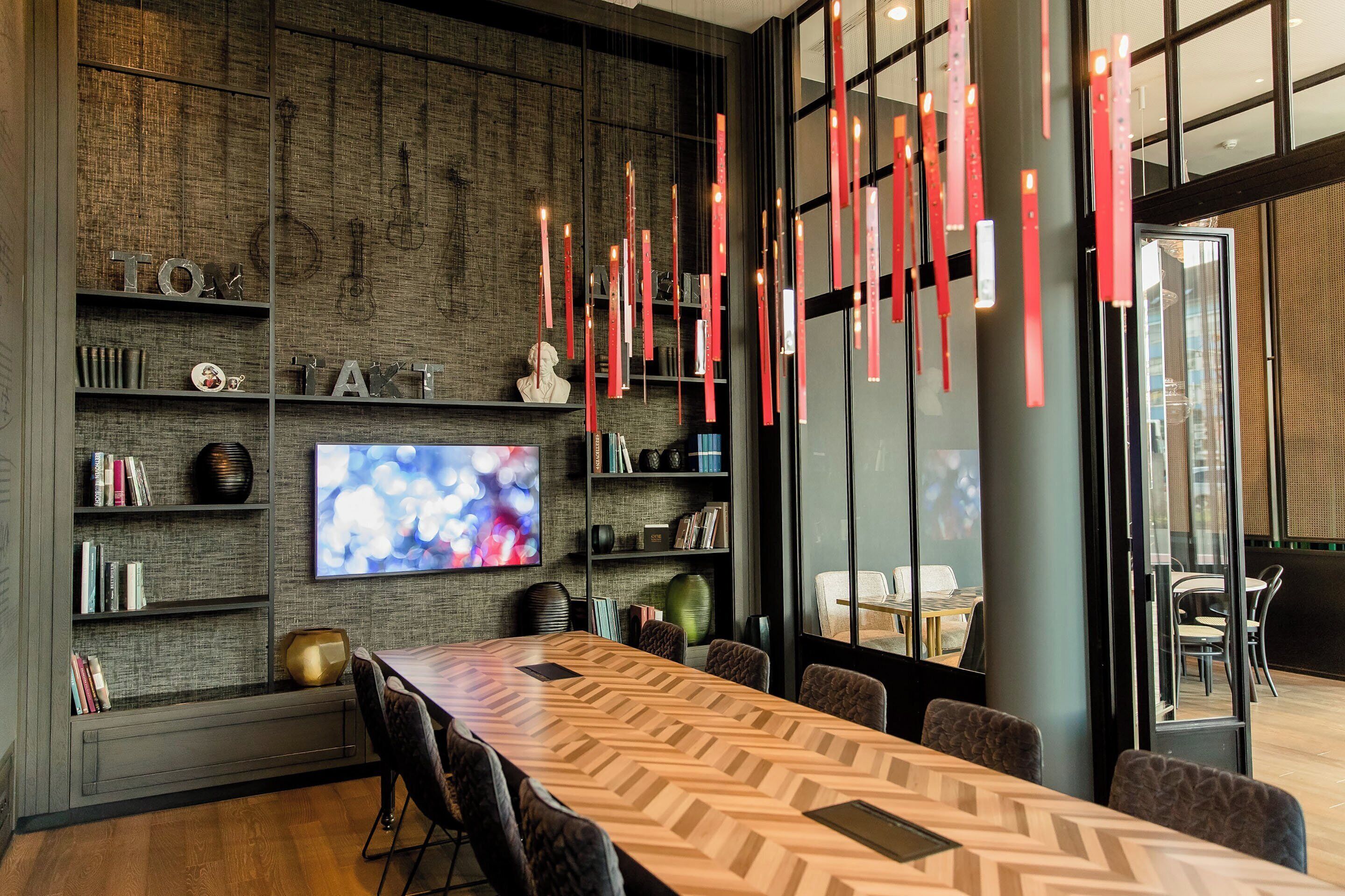 A modern conference room with a large wooden table, comfortable seating, a flat-screen TV, and decorative red elements.