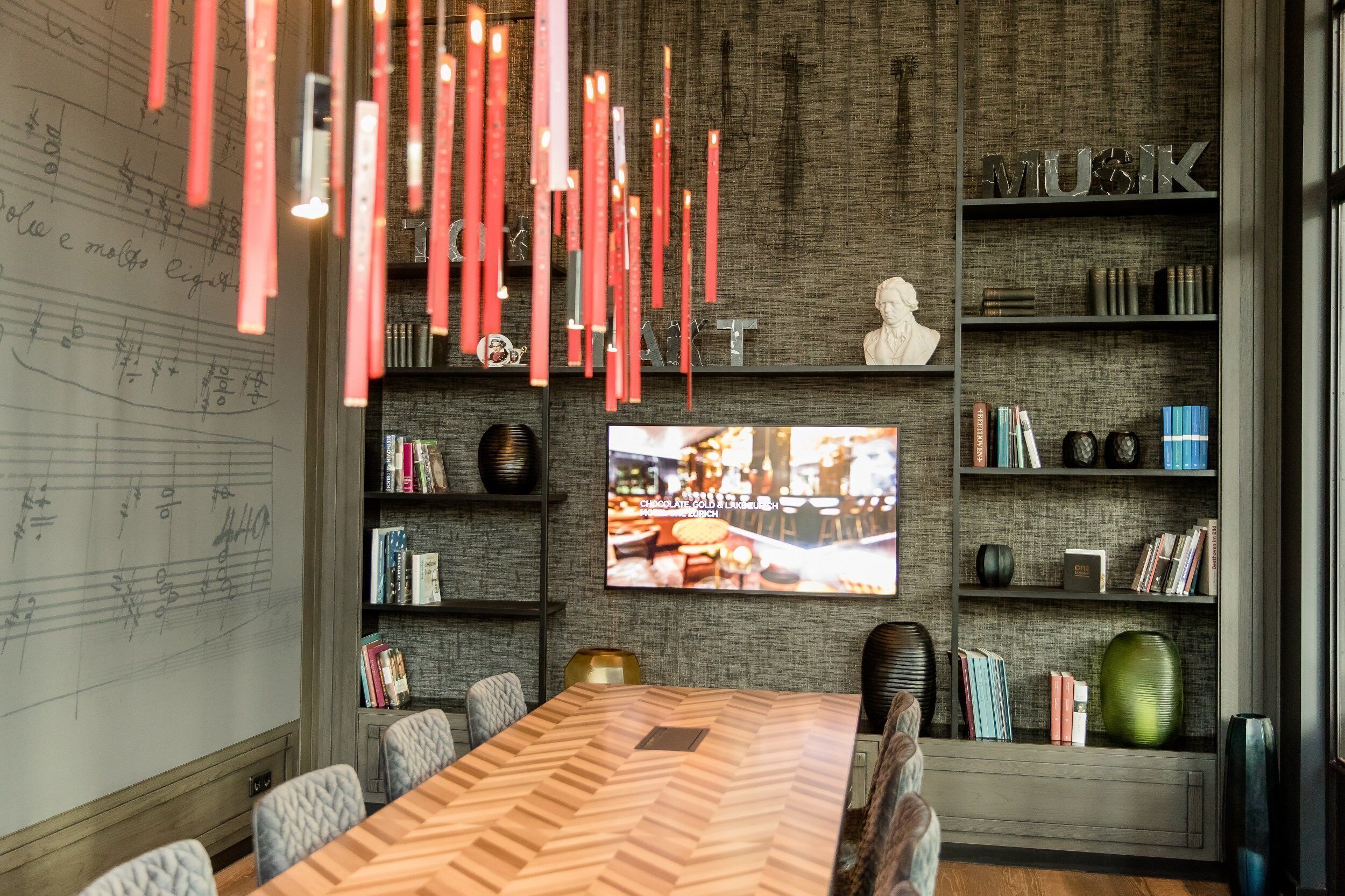 Una sala conferenze moderna con un ampio tavolo di legno, sedute confortevoli, una lavagna con note musicali, uno schermo televisivo e scaffali contenenti diversi oggetti.
