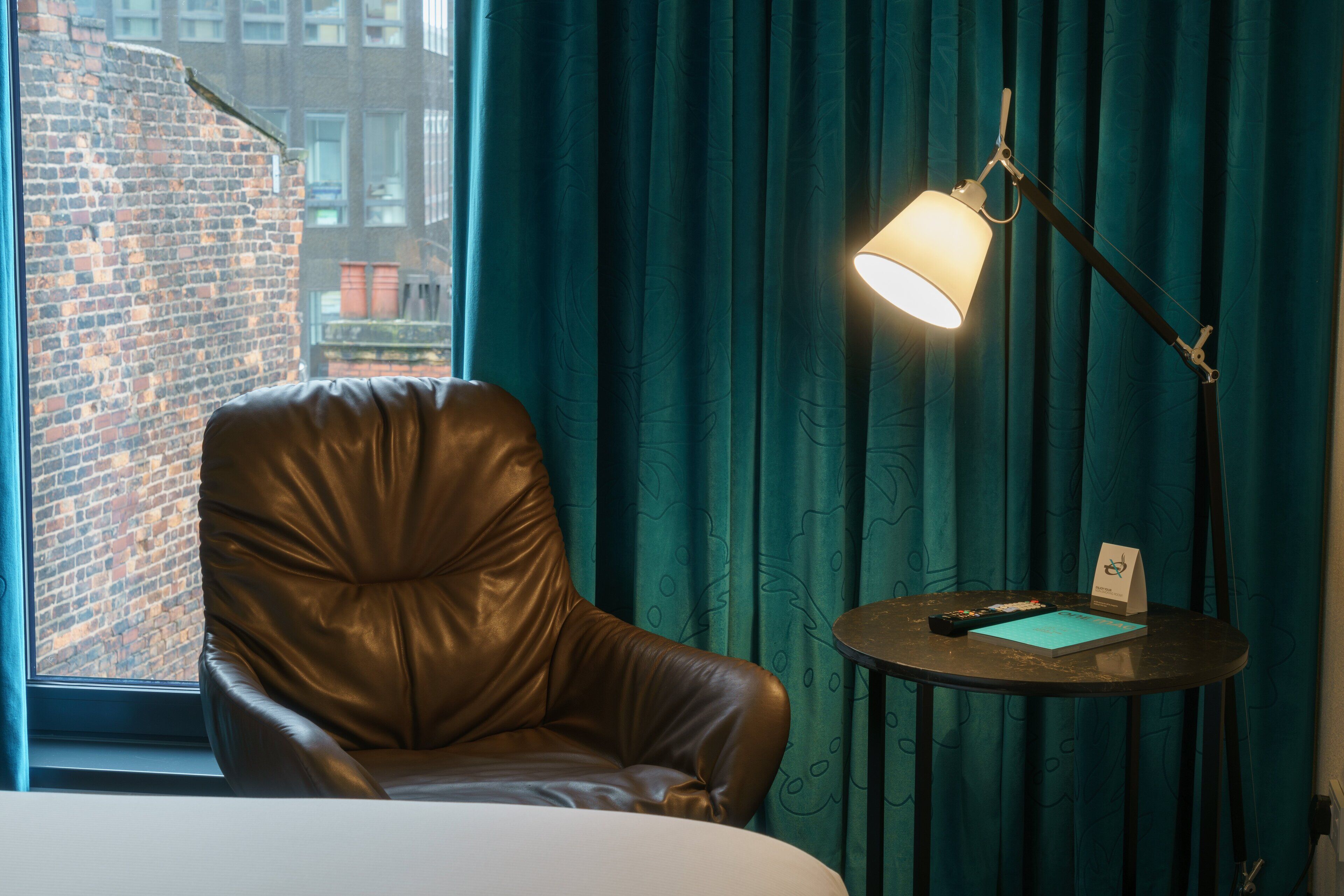 A hotel room with a leather chair, a bedside table with a lamp, a remote control, and a notebook.