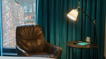 A hotel room with a leather chair, a bedside table with a lamp, a remote control, and a notebook.