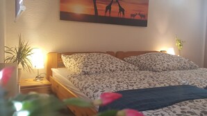 A bedroom with a bed, two bedside lamps, a nightstand, a potted plant, and a wall-mounted painting of a savannah scene. - Ferienhaus am Schloss Sayn (Bendorf)