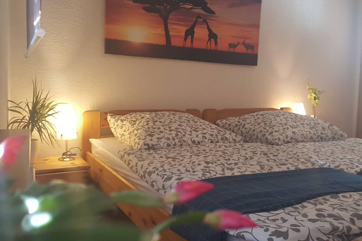Un dormitorio con una cama, dos lámparas de noche, una mesita de noche, una planta en maceta y un cuadro colgado en la pared que muestra una escena de sabana.