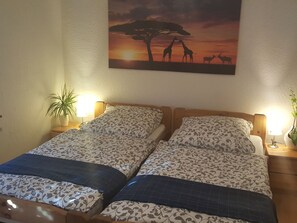 Un lit double avec une literie à motifs, deux lampes de chevet, un tableau représentant un paysage de savane accroché au mur et une plante en pot sur le côté gauche.