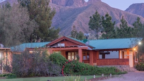 A wooden cabin with a green roof, surrounded by lush greenery and mountains in the background. - Entre Montañas y Rio (Cacheuta)