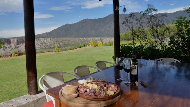 A table set outdoors with a view of mountains, a wooden board with various cheeses and meats, a bottle of wine, and glasses. - Entre Montañas y Rio (Cacheuta)