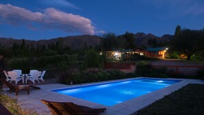 A swimming pool with a wooden deck and white chairs, surrounded by greenery and mountains in the background. - Entre Montañas y Rio (Cacheuta)