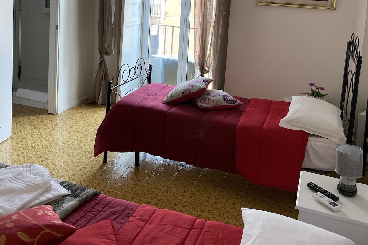 A hotel room with two beds, a shower, a painting on the wall, and a view through the window.