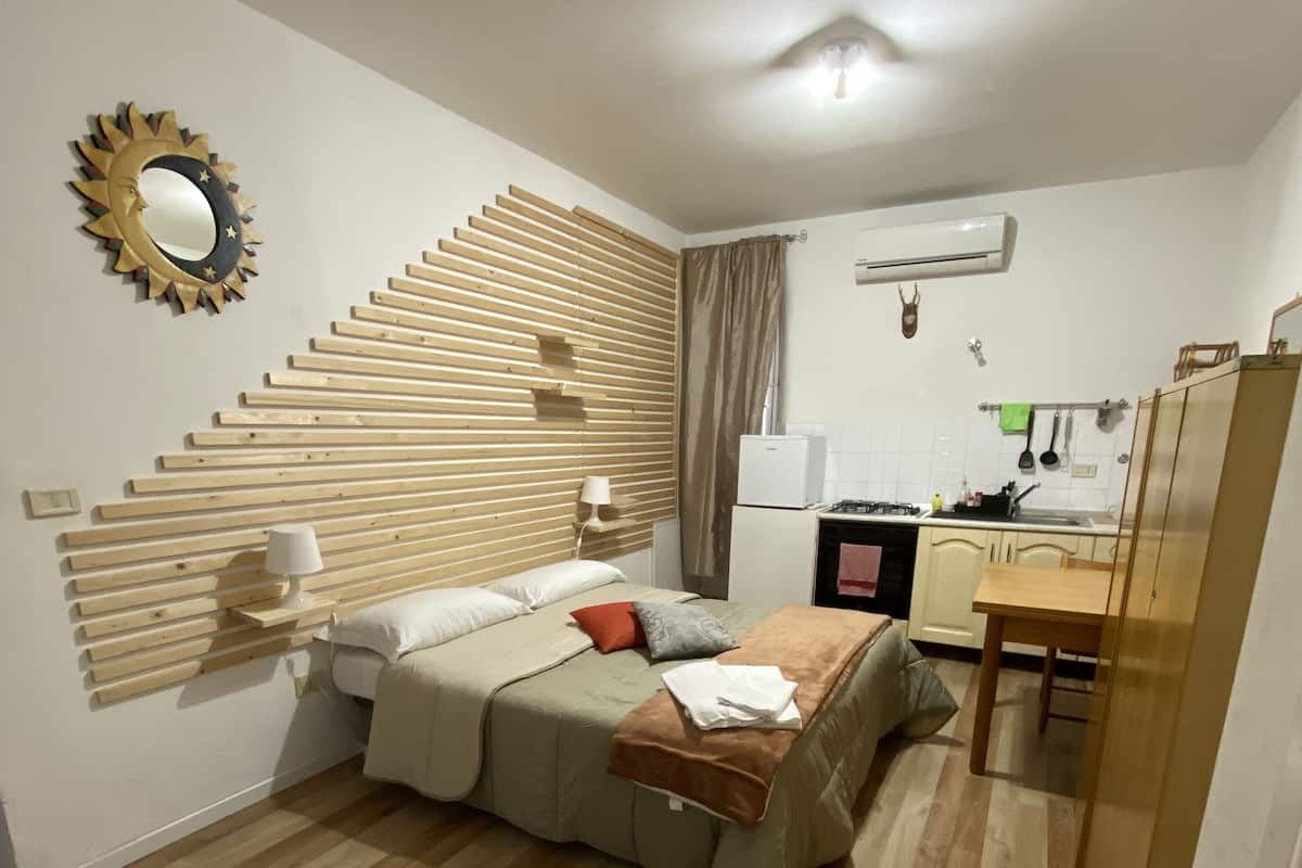 A compact hotel room with a bed, a small kitchenette, a wooden floor, and a decorative wall panel.