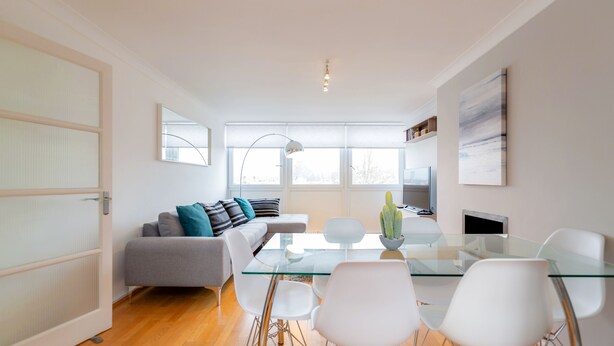 Un salon moderne avec une table à manger en verre, des chaises blanches, un canapé gris agrémenté de coussins bleus, une plante en pot (un cactus) et une grande fenêtre offrant une vue dégagée.