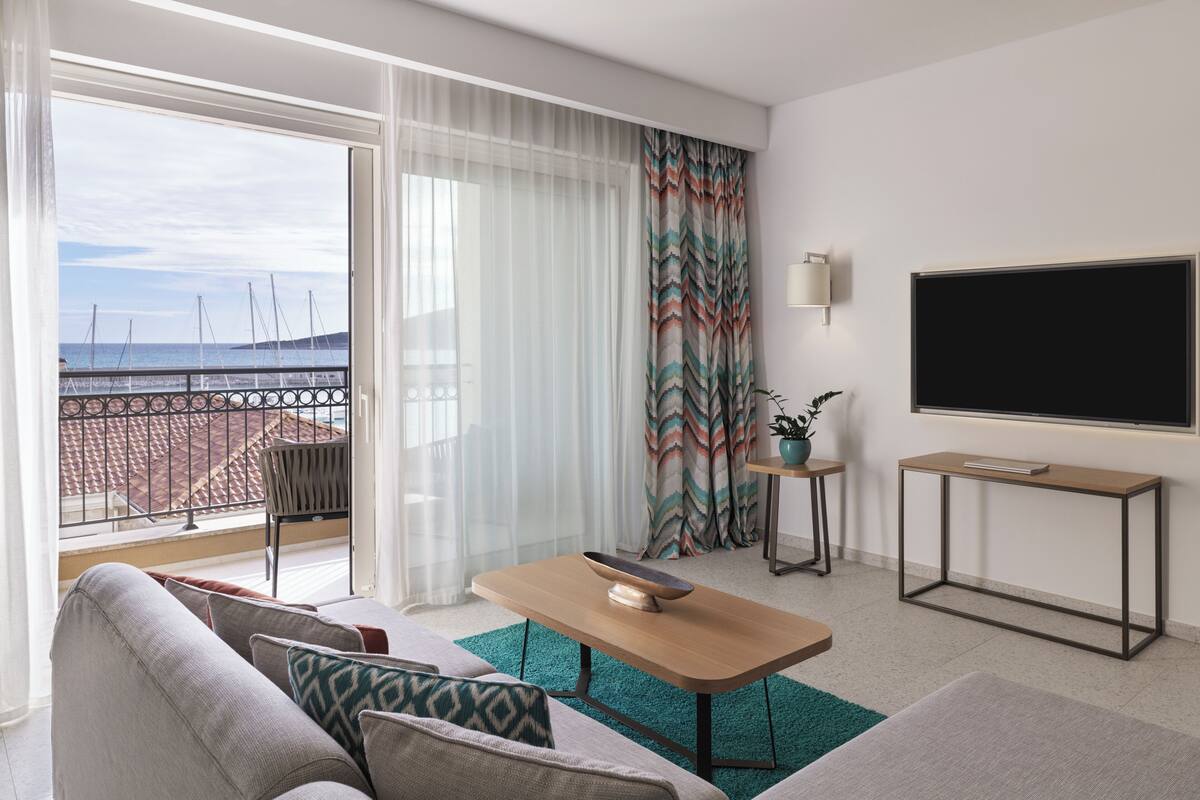 A modern hotel room with a balcony, a large TV, a small table, and a sofa.