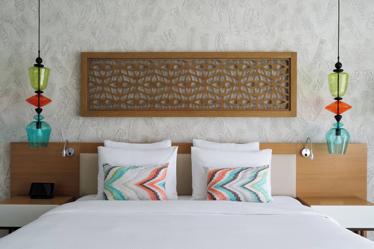 A hotel room with a bed, two bedside tables, a nightstand, a decorative wall panel, and two hanging lamps.