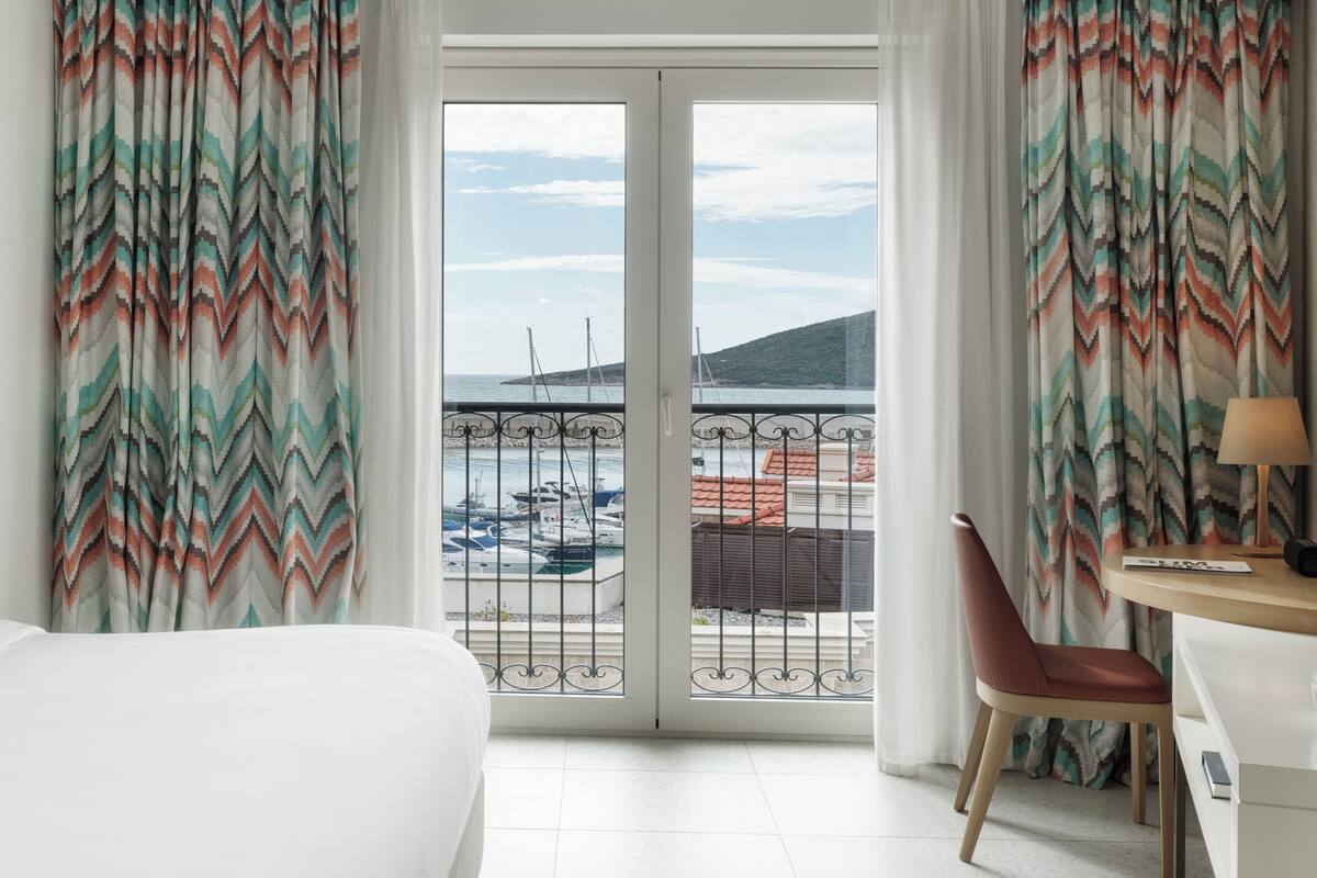 A hotel room with a bed, a desk, a chair, and a view of boats and a hilly landscape through the balcony doors.