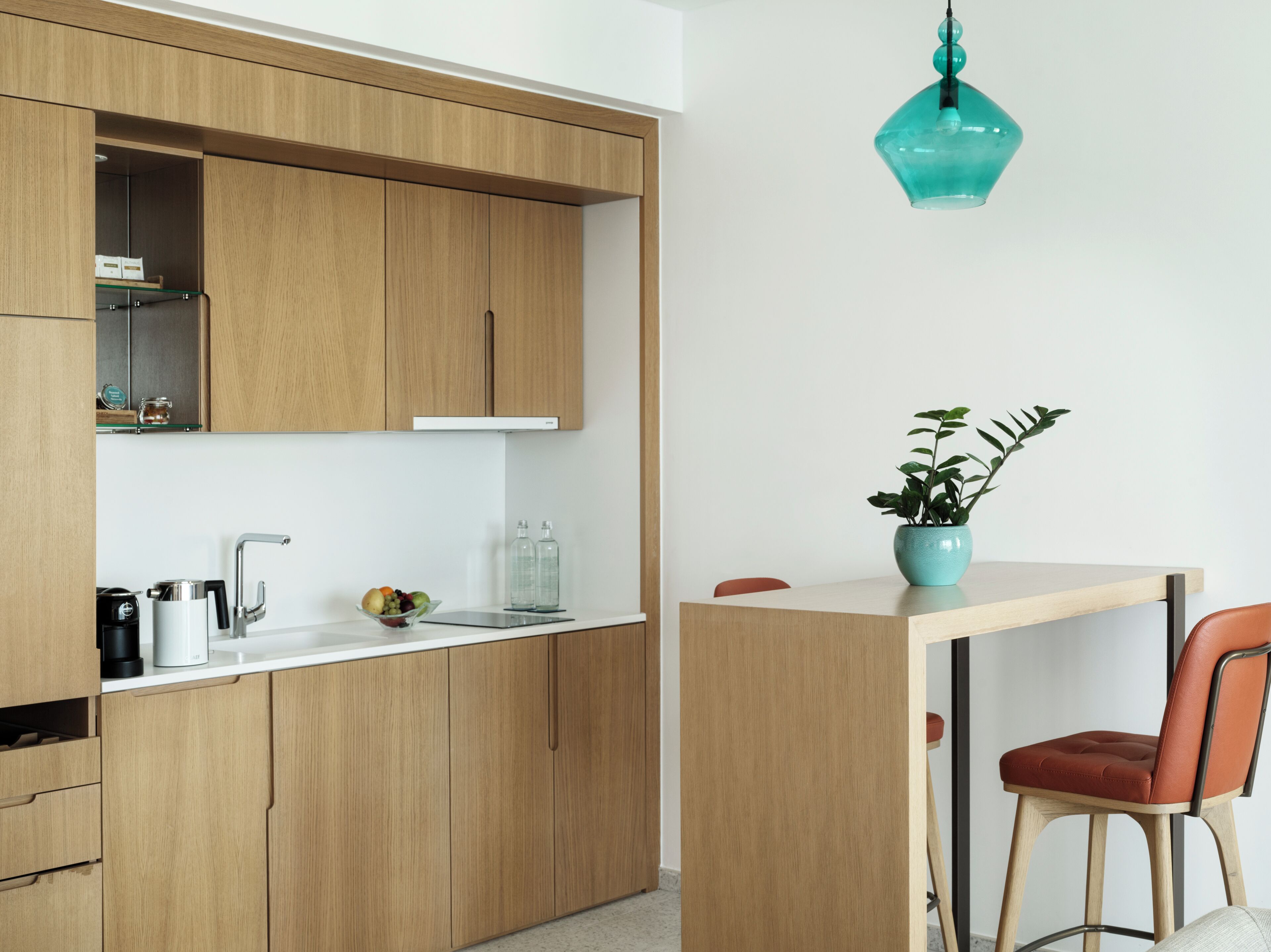 A modern kitchen with wooden cabinets, a white countertop, a coffee machine, a fruit bowl, and a teal pendant light.