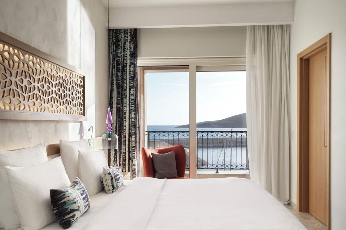 A hotel room with a large bed, a decorative headboard, a view of the sea, and a balcony with a railing.