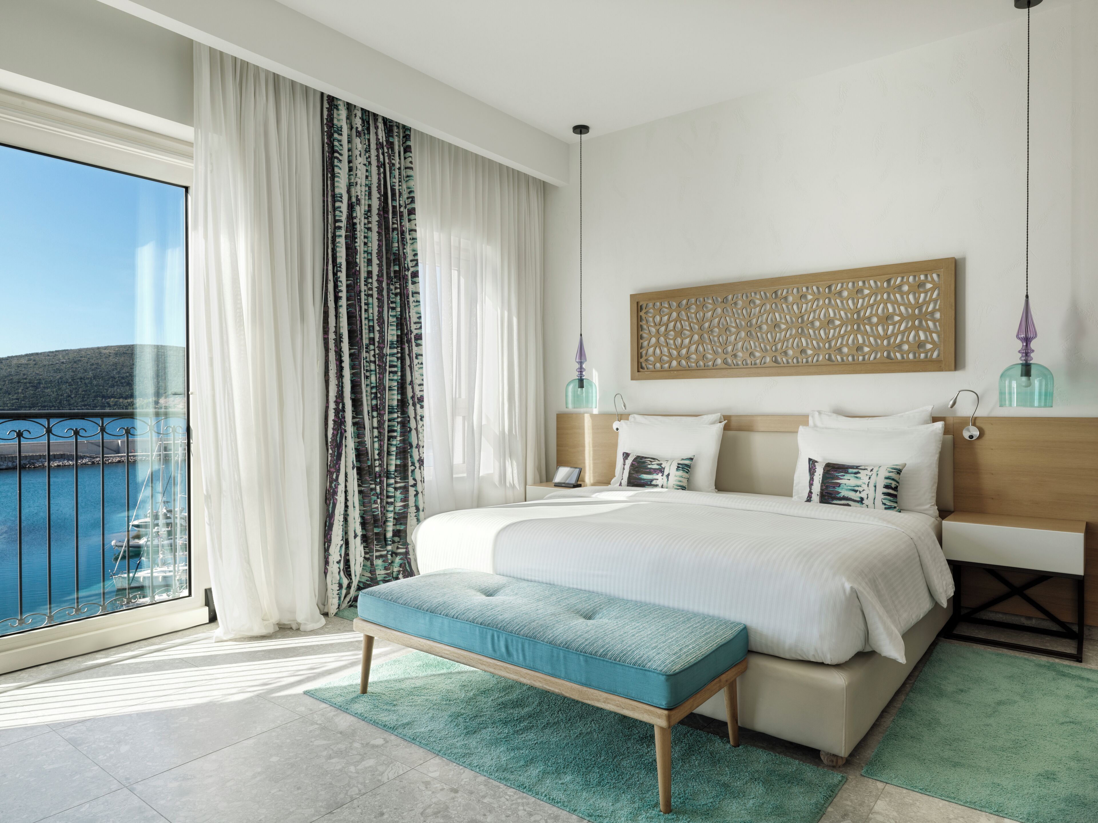 A modern hotel room with a large bed, a bench, a view of the sea, and a decorative wall panel.