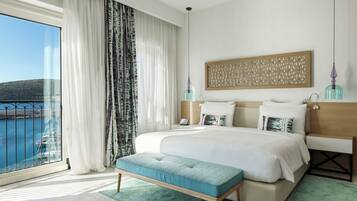 A modern hotel room with a large bed, a bench, a view of the sea, and a decorative wall panel.
