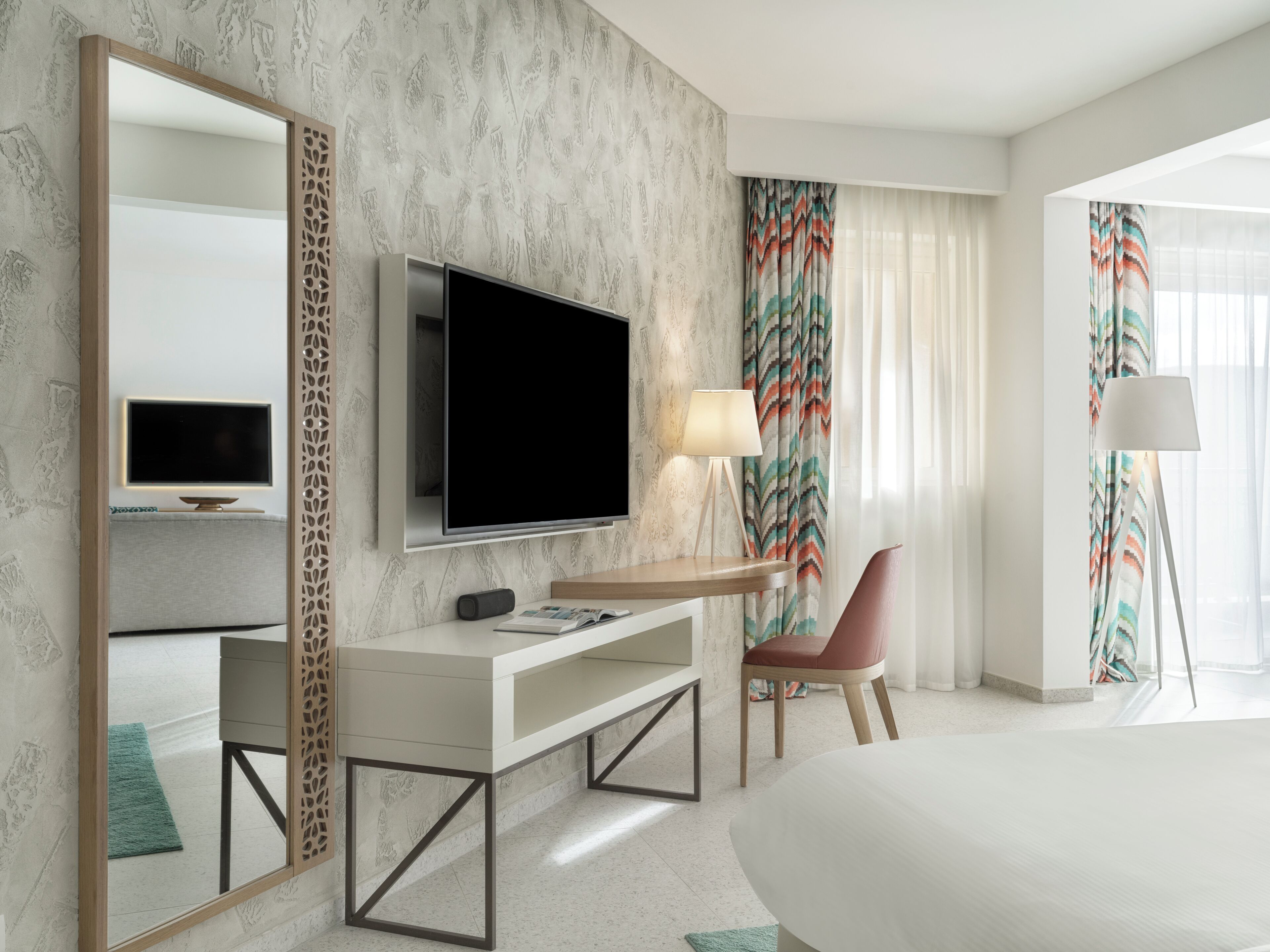A modern hotel room with a large bed, a TV mounted on the wall, a desk with a lamp, and a chair.