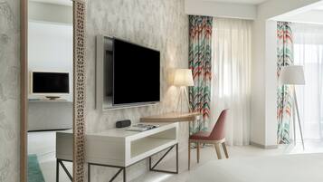 A modern hotel room with a large bed, a TV mounted on the wall, a desk with a lamp, and a chair.