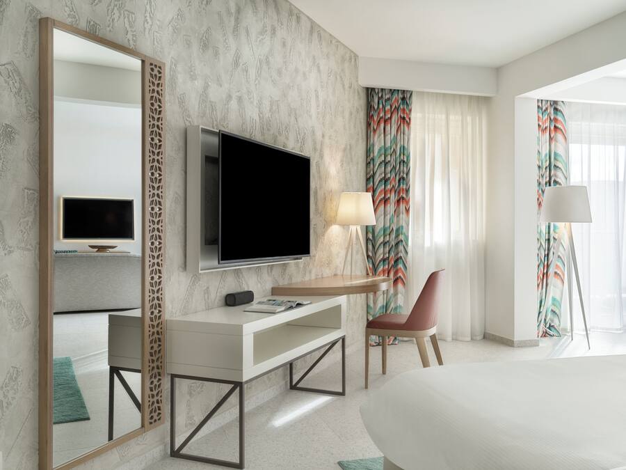A modern hotel room with a large bed, a TV mounted on the wall, a desk with a lamp, and a chair.