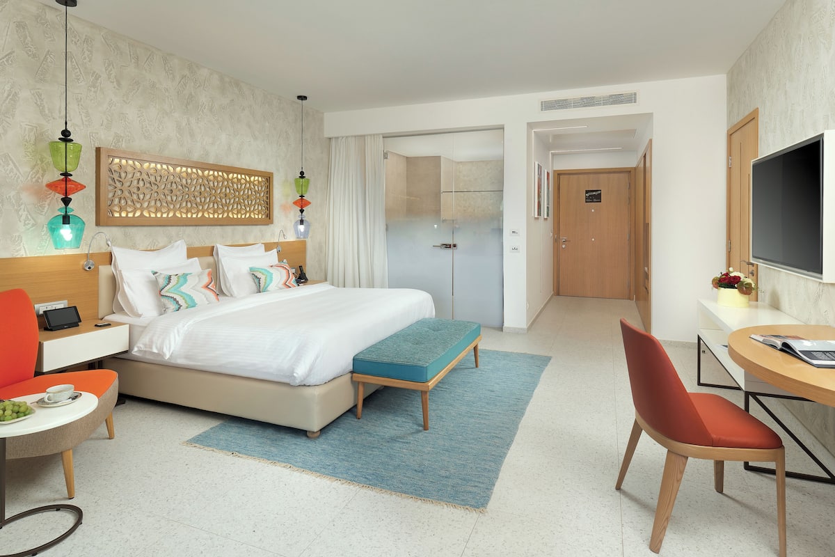 A modern hotel room with a bed, bedside table, chair, desk, and TV.