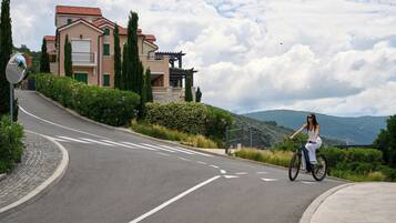 A curved road with a cyclist, a large house with a balcony, and a mountainous backdrop.
