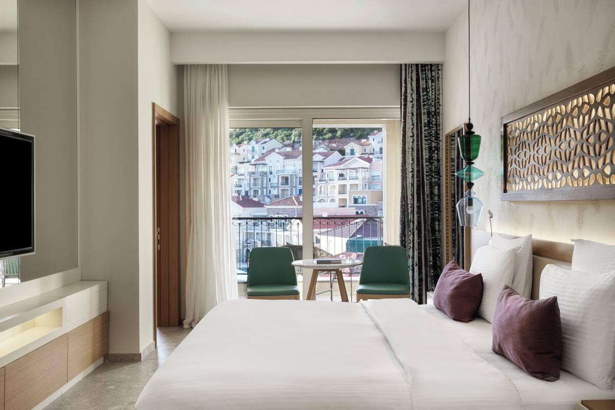 A hotel room with a bed, a TV, a balcony with a view of buildings, and a dining area with chairs.