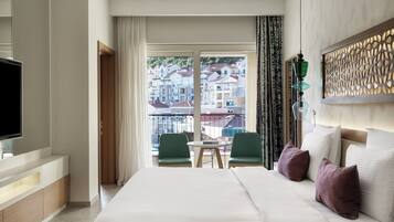 Ein Hotelzimmer mit Bett, Fernseher, einem Balkon mit Blick auf Gebäude und einer Essecke mit Stühlen.
