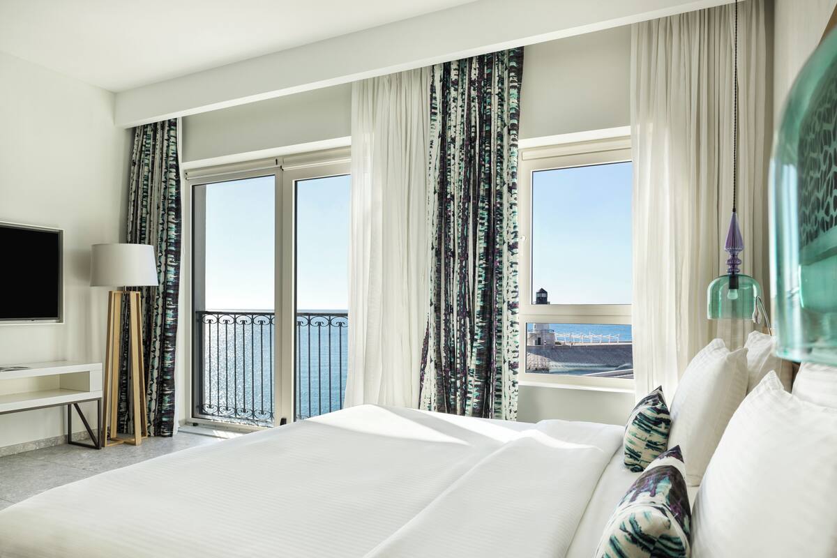 A hotel room with a large bed, a TV, a desk, and a view of the sea.