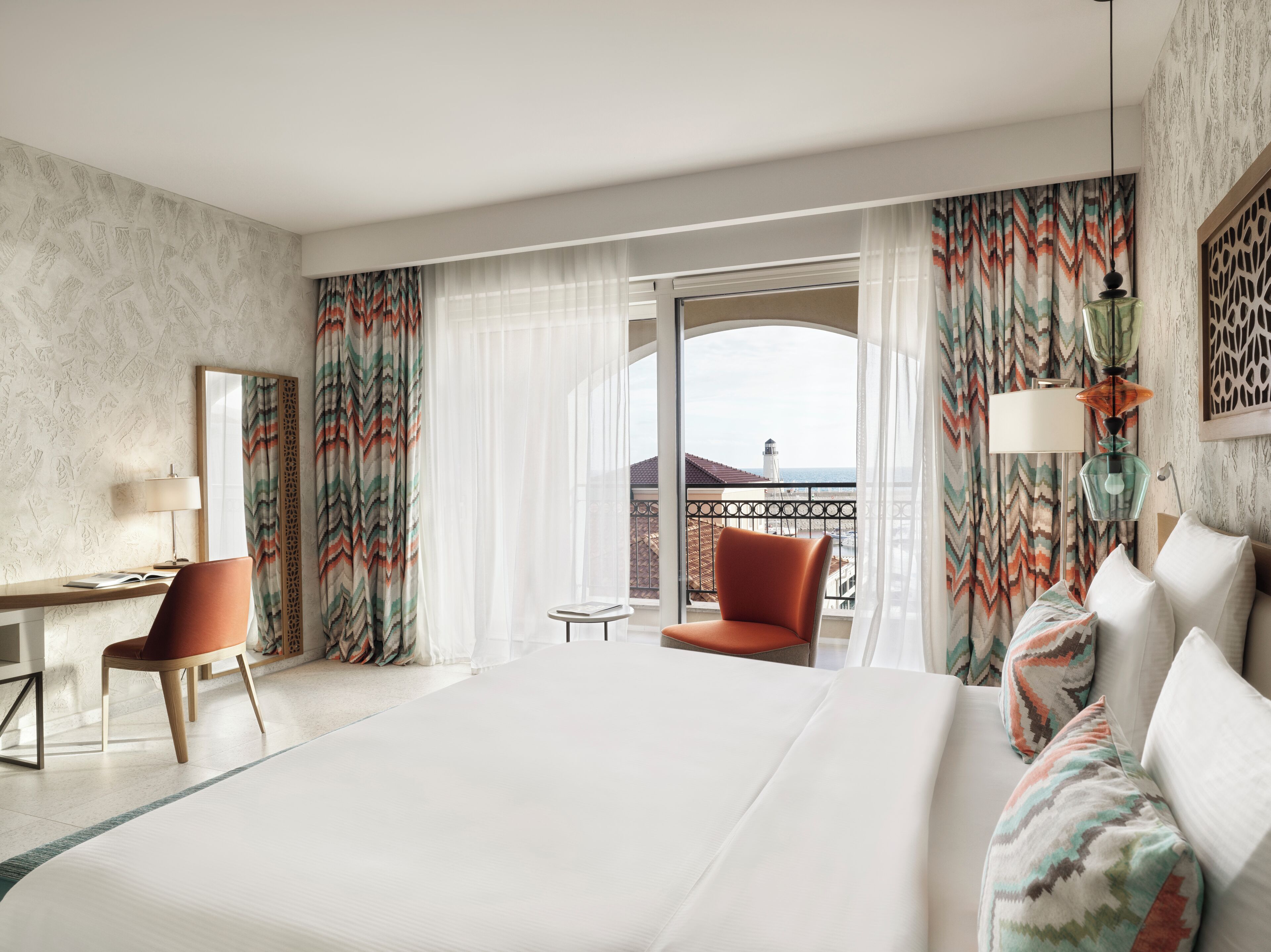 A hotel room with a large bed, a desk, a chair, and a balcony with a view of the city.