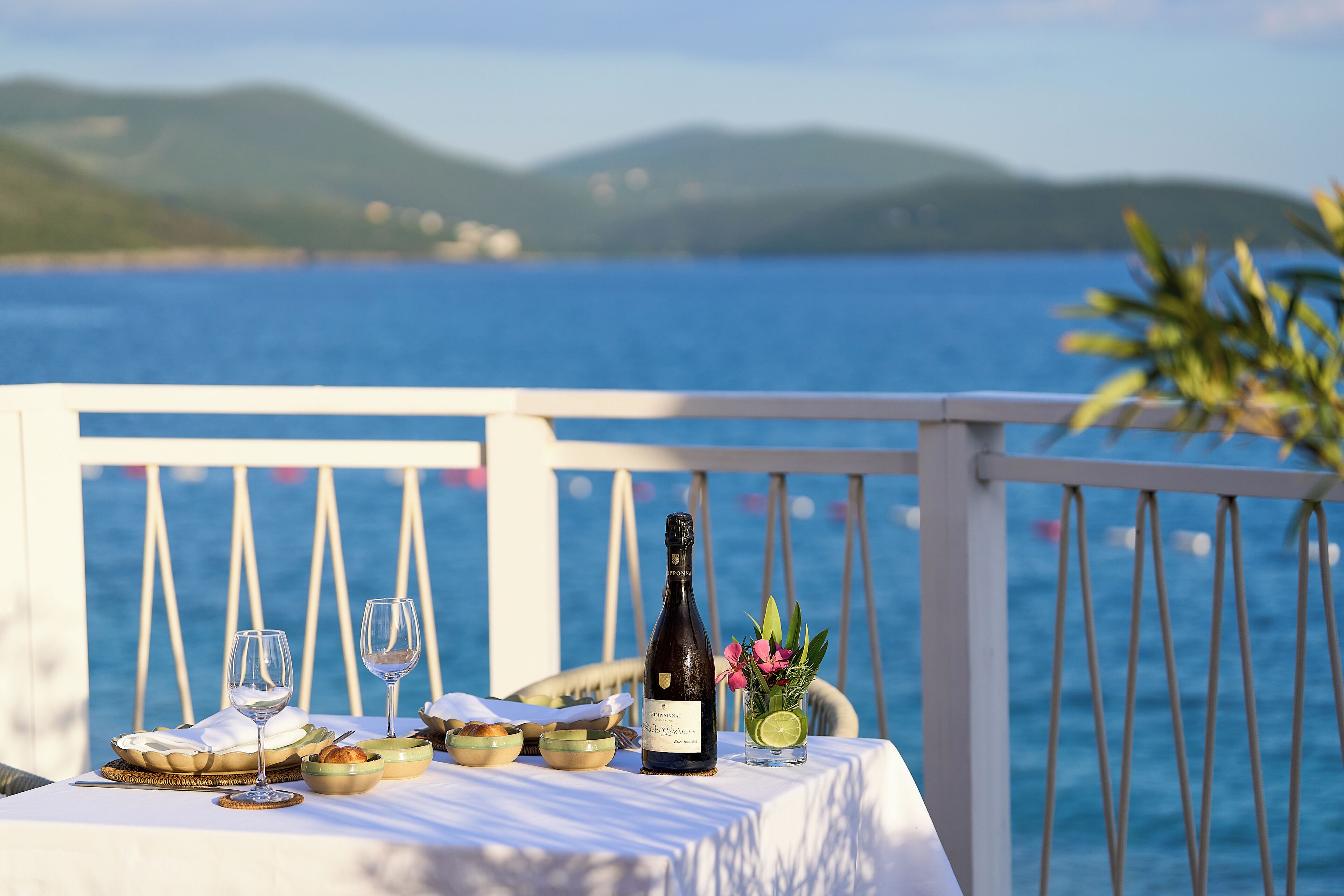 A table set with a bottle of wine, glasses, and a glass of water with a slice of lime, on a balcony with a view of the sea and hills.