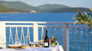 A table set with a bottle of wine, glasses, and a glass of water with a slice of lime, on a balcony with a view of the sea and hills.