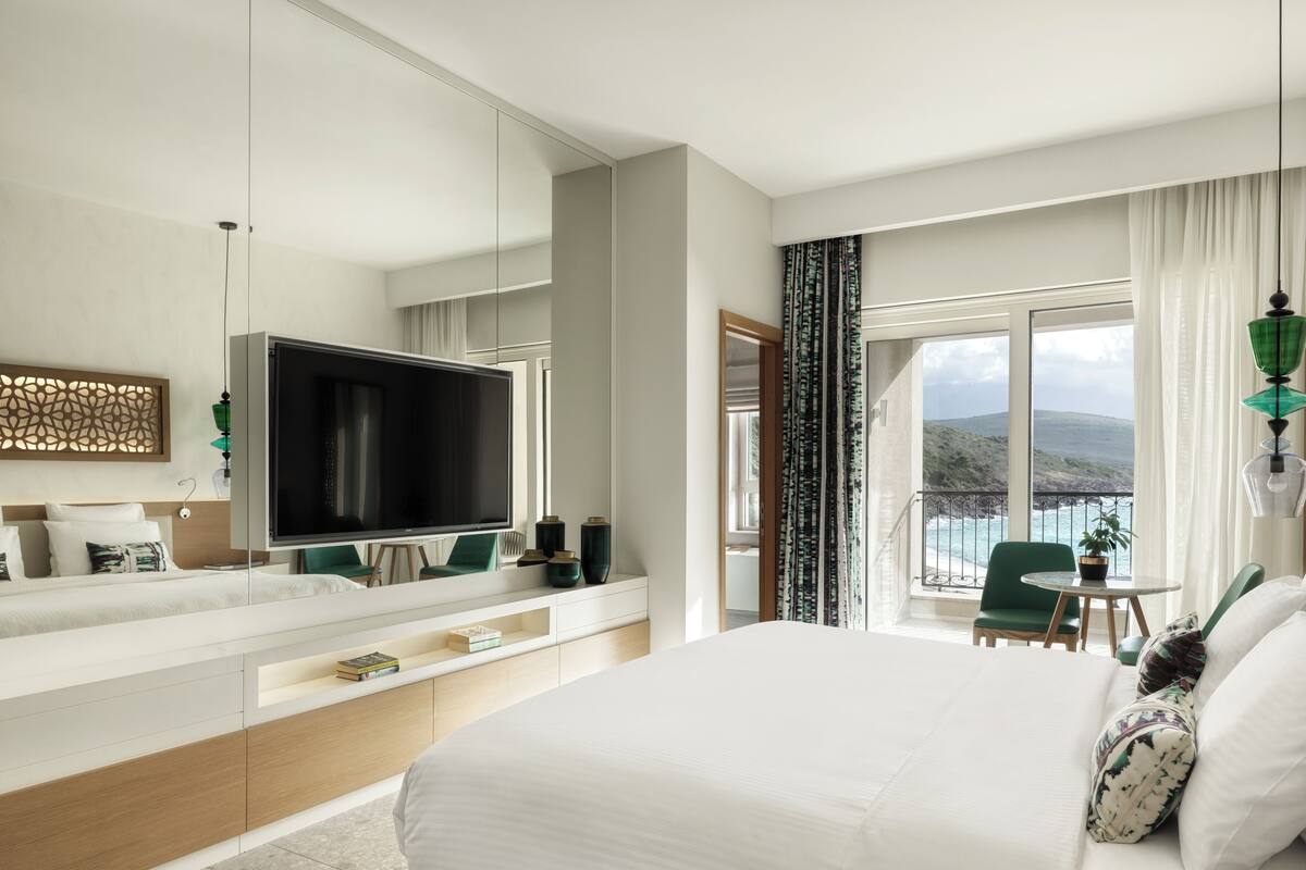 A modern hotel room with a large bed, a TV, a balcony with a view of the sea, and a dining area.