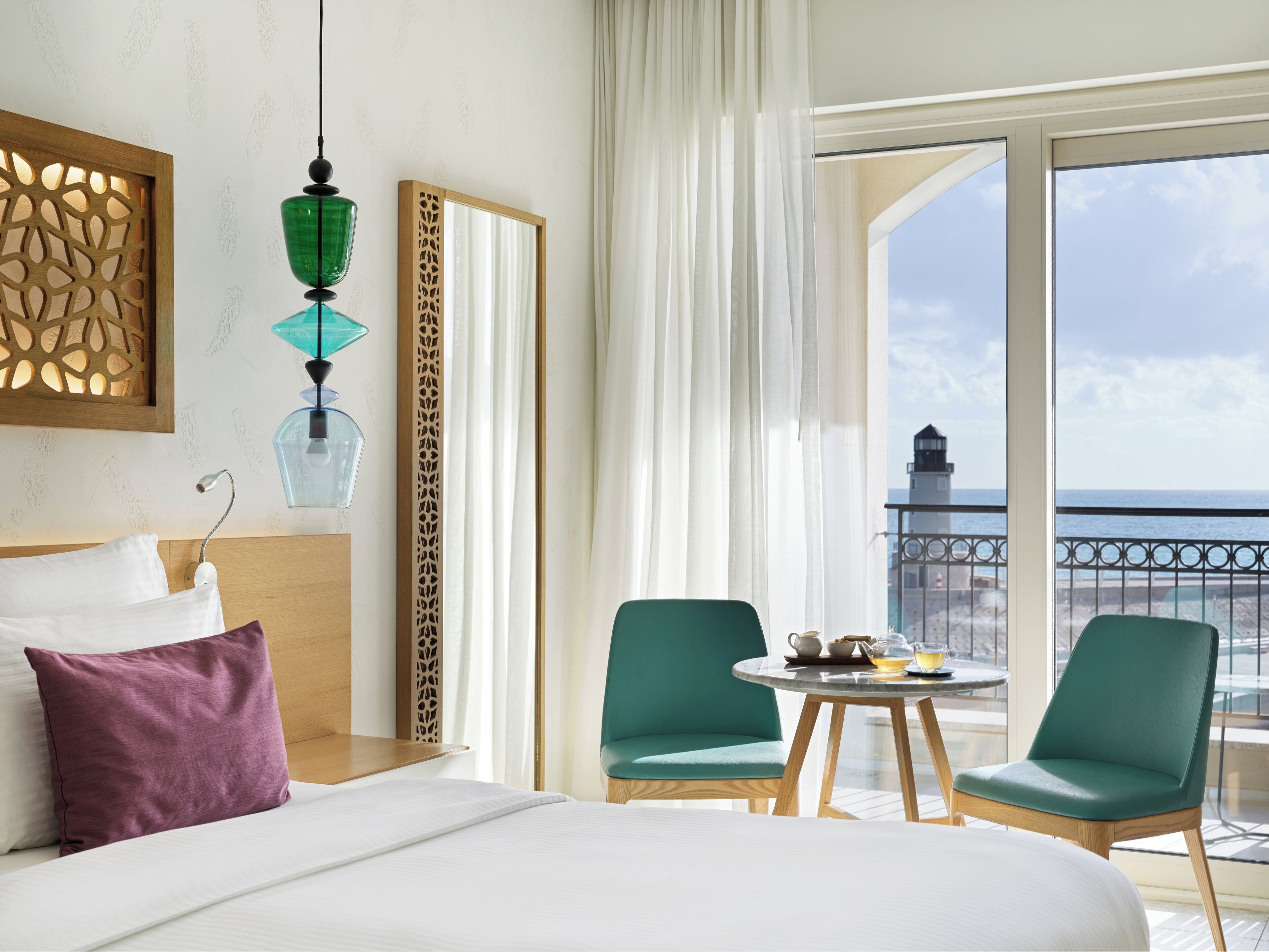 A hotel room with a bed, two chairs, a small table with tea set, and a view of the sea and a lighthouse.