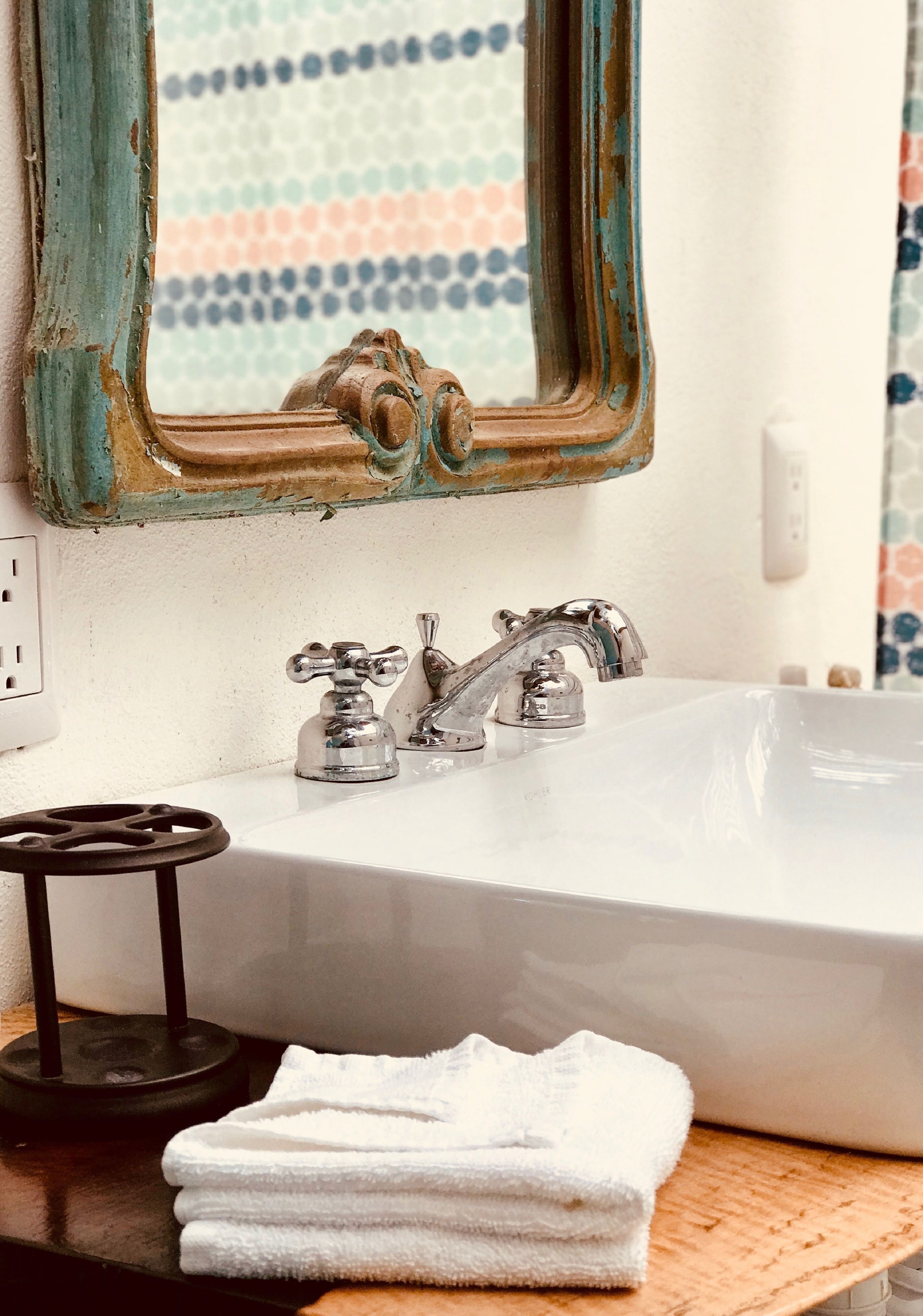 Une salle de bain avec un lavabo blanc, deux robinets, un miroir au cadre vieilli et une serviette posée sur le comptoir.