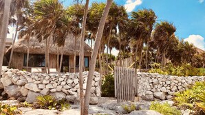 Una estructura con techo de paja y un muro de piedra frontal, rodeada de palmeras y vegetación tropical.