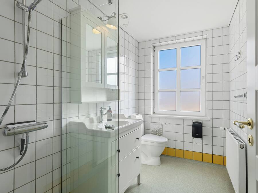Un baño con cabina de ducha de vidrio, lavamanos blanco, inodoro y ventana con vistas.