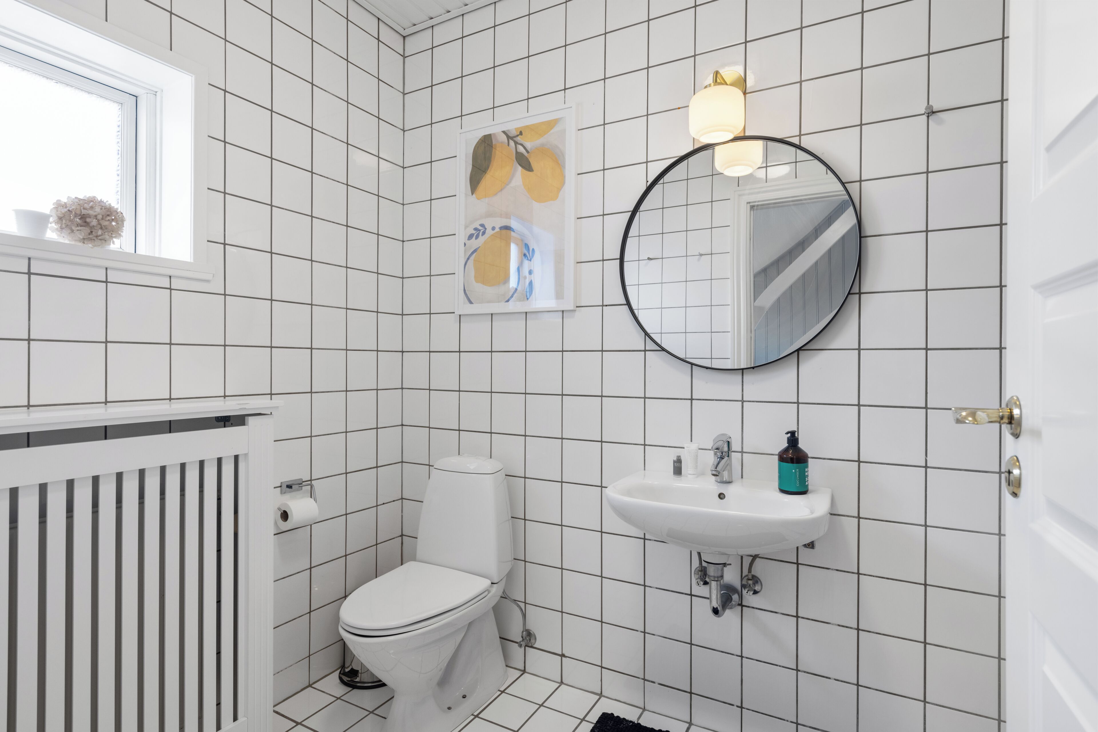 Un baño con paredes de azulejos blancos, un inodoro, un lavabo, un espejo y una ventana con una planta.