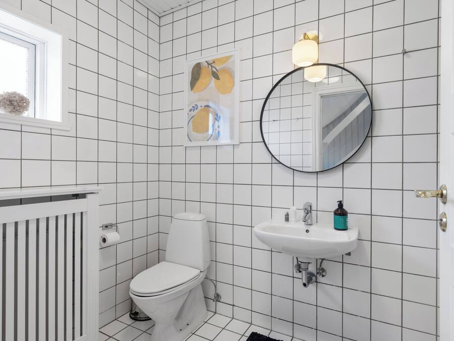 A bathroom with white tiled walls, a toilet, a sink, a mirror, and a window with a plant.