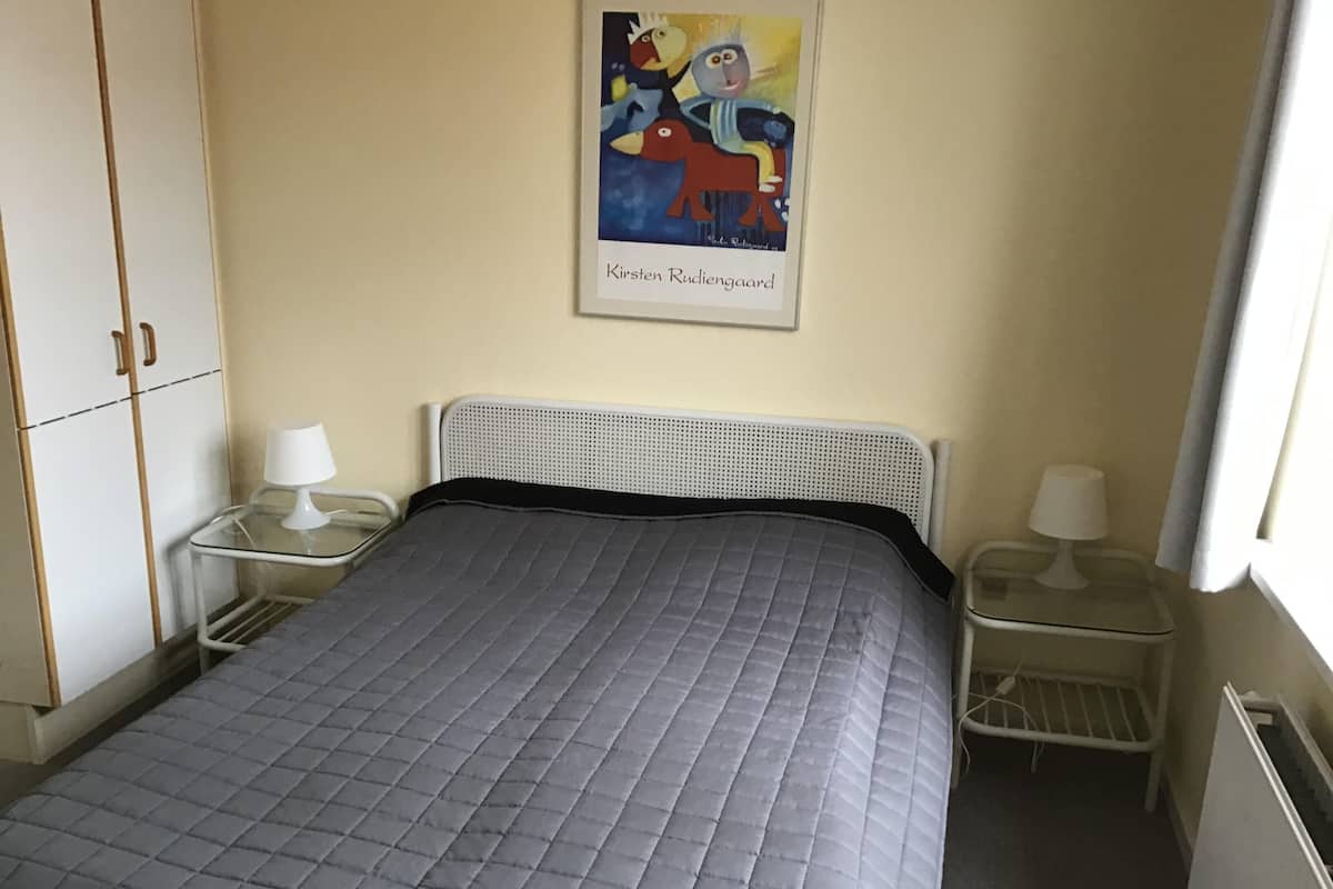 A neatly made bed with a grey quilt, two bedside tables with lamps, a framed picture on the wall, and a wardrobe.