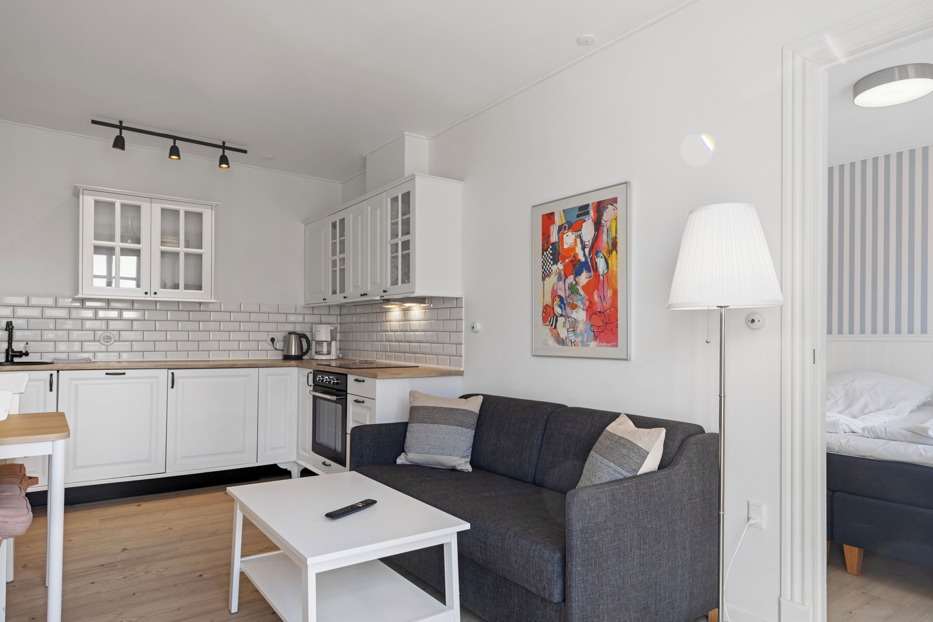 Et moderne køkken med hvide skabe, en kaffemaskine og en elkedel. En grå sofa med en fjernbetjening står på et hvidt bord. Et farverigt abstrakt maleri hænger på væggen.
