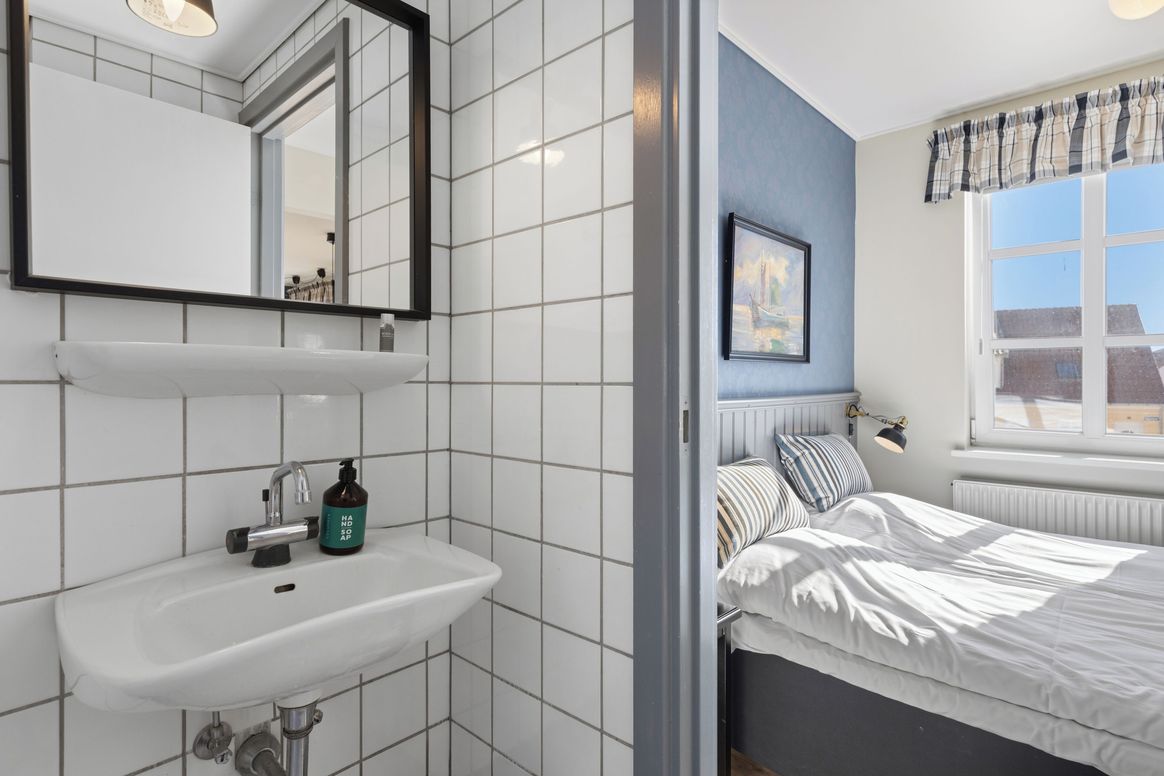 Un baño con lavamanos blanco, espejo y ventana con vistas a edificios.