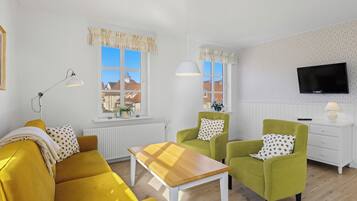 Una habitación con un sofá amarillo, dos sillones verdes, una mesa de centro de madera, un aparador blanco, un televisor y una ventana con cortinas.
